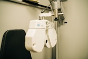 A close-up view of an ophthalmology machine, typically used for eye examinations. The device features a white, industrial design with various lenses and adjustment knobs. It is mounted on a metallic arm attached to a support stand, next to a black cushioned chair.