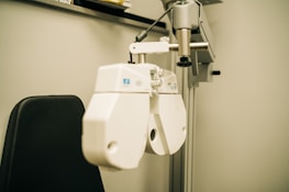 A close-up view of an ophthalmology machine, typically used for eye examinations. The device features a white, industrial design with various lenses and adjustment knobs. It is mounted on a metallic arm attached to a support stand, next to a black cushioned chair.