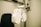 A close-up view of an ophthalmology machine, typically used for eye examinations. The device features a white, industrial design with various lenses and adjustment knobs. It is mounted on a metallic arm attached to a support stand, next to a black cushioned chair.