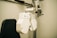 A close-up view of an ophthalmology machine, typically used for eye examinations. The device features a white, industrial design with various lenses and adjustment knobs. It is mounted on a metallic arm attached to a support stand, next to a black cushioned chair.