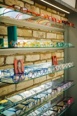 Several shelves display a variety of colorful eyeglasses against a brick wall, with each shelf showcasing different colors such as red, orange, yellow, blue, and purple. Small portrait photos are placed intermittently among the glasses.
