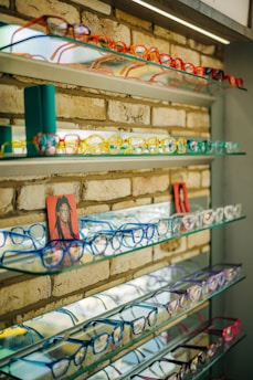 Several shelves display a variety of colorful eyeglasses against a brick wall, with each shelf showcasing different colors such as red, orange, yellow, blue, and purple. Small portrait photos are placed intermittently among the glasses.