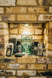 A collection of vintage mobile phones is arranged around a transparent glass head with glasses perched on it. The setting is a brick alcove with a small light above, casting a warm glow over the scene.
