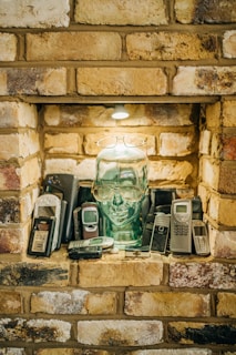 A collection of vintage mobile phones is arranged around a transparent glass head with glasses perched on it. The setting is a brick alcove with a small light above, casting a warm glow over the scene.