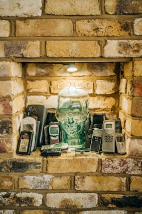 A collection of vintage mobile phones is arranged around a transparent glass head with glasses perched on it. The setting is a brick alcove with a small light above, casting a warm glow over the scene.