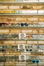 Multiple shelves filled with a variety of eyeglasses are mounted on a brick wall. The glasses are arranged neatly, showcasing different styles and colors, including frames in yellow, green, blue, and black. A small sign reading 'EYES' is placed on some of the shelves.