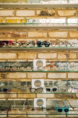 Multiple shelves filled with a variety of eyeglasses are mounted on a brick wall. The glasses are arranged neatly, showcasing different styles and colors, including frames in yellow, green, blue, and black. A small sign reading 'EYES' is placed on some of the shelves.