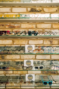 Multiple shelves filled with a variety of eyeglasses are mounted on a brick wall. The glasses are arranged neatly, showcasing different styles and colors, including frames in yellow, green, blue, and black. A small sign reading 'EYES' is placed on some of the shelves.