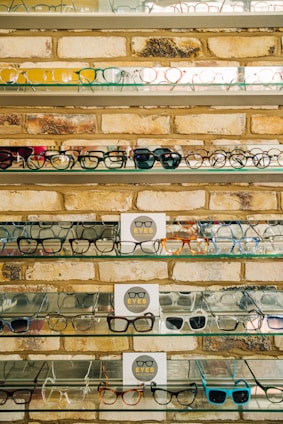 Multiple shelves filled with a variety of eyeglasses are mounted on a brick wall. The glasses are arranged neatly, showcasing different styles and colors, including frames in yellow, green, blue, and black. A small sign reading 'EYES' is placed on some of the shelves.