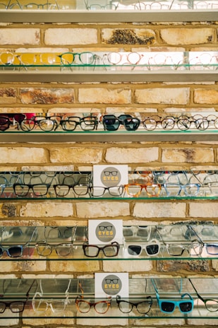 Multiple shelves filled with a variety of eyeglasses are mounted on a brick wall. The glasses are arranged neatly, showcasing different styles and colors, including frames in yellow, green, blue, and black. A small sign reading 'EYES' is placed on some of the shelves.