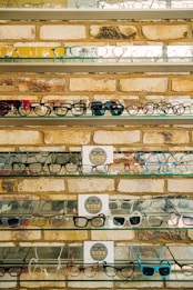 Multiple shelves filled with a variety of eyeglasses are mounted on a brick wall. The glasses are arranged neatly, showcasing different styles and colors, including frames in yellow, green, blue, and black. A small sign reading 'EYES' is placed on some of the shelves.