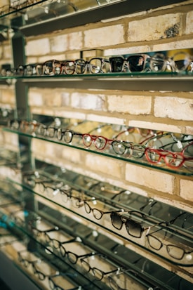 Multiple pairs of eyeglasses are neatly displayed on glass shelves against a brick wall. The glasses vary in style, shape, and color, creating a visually appealing array.
