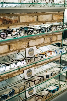 A display of various eyeglasses is arranged neatly on shelves set against a brick wall. The shelves are made of glass, reflecting light and displaying multiple styles and colors of frames. There are informational cards placed among the glasses featuring the word 'EYES' alongside a simple graphic of eyeglasses.
