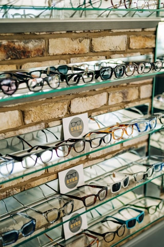 A display of various eyeglasses is arranged neatly on shelves set against a brick wall. The shelves are made of glass, reflecting light and displaying multiple styles and colors of frames. There are informational cards placed among the glasses featuring the word 'EYES' alongside a simple graphic of eyeglasses.