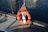 An orange lifebuoy with reflective strips is attached to a metal railing by an orange rope. It is situated on a wooden dock next to calm, rippling water.