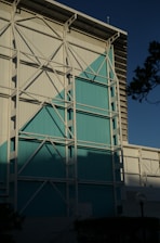 A large industrial-style building facade featuring white and teal panels. The structure is reinforced with a visible white metal framework. The scene is set against a clear sky, casting shadows on the building, with some greenery and a lamp post visible in the foreground.