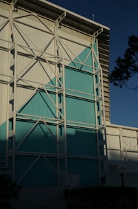A large industrial-style building facade featuring white and teal panels. The structure is reinforced with a visible white metal framework. The scene is set against a clear sky, casting shadows on the building, with some greenery and a lamp post visible in the foreground.