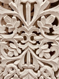 Intricate wooden carving featuring elaborate floral and leaf patterns, with a symmetrical design. The craftsmanship showcases detailed scrollwork and a textured surface.