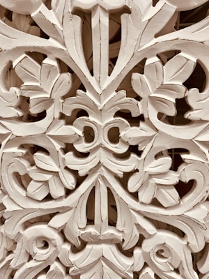 Intricate wooden carving featuring elaborate floral and leaf patterns, with a symmetrical design. The craftsmanship showcases detailed scrollwork and a textured surface.