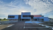 A large, modern healthcare facility stands under a partly cloudy sky with the sun peeking out from behind. The building has a mix of white, gray, and pastel pink sections and features the signage 'Klinik Kesihatan Bandaraya Miri'. The foreground shows an empty parking lot with clear markings and surrounding greenery.