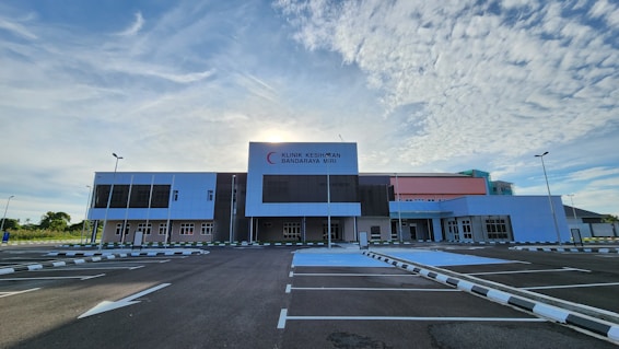 A large, modern healthcare facility stands under a partly cloudy sky with the sun peeking out from behind. The building has a mix of white, gray, and pastel pink sections and features the signage 'Klinik Kesihatan Bandaraya Miri'. The foreground shows an empty parking lot with clear markings and surrounding greenery.