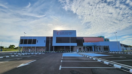 A large, modern healthcare facility stands under a partly cloudy sky with the sun peeking out from behind. The building has a mix of white, gray, and pastel pink sections and features the signage 'Klinik Kesihatan Bandaraya Miri'. The foreground shows an empty parking lot with clear markings and surrounding greenery.