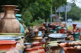 A variety of large, decorative ceramic vases are displayed along the side of a road. The vases feature different designs and colors, including earthy browns, vibrant blues, and muted greens. They are stacked closely together, creating an artistic and colorful arrangement. In the background, a road with occasional vehicles and surrounding greenery is visible.