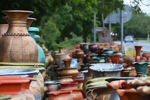 A variety of large, decorative ceramic vases are displayed along the side of a road. The vases feature different designs and colors, including earthy browns, vibrant blues, and muted greens. They are stacked closely together, creating an artistic and colorful arrangement. In the background, a road with occasional vehicles and surrounding greenery is visible.