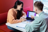 Two people are sitting at a table with a laptop, engaged in a discussion. The person on the right is pointing at the laptop screen, which displays some data or website interface. They are in a modern, well-lit environment with a red cushioned bench.