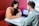 Two people are sitting at a table with a laptop, engaged in a discussion. The person on the right is pointing at the laptop screen, which displays some data or website interface. They are in a modern, well-lit environment with a red cushioned bench.