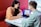 Two people are sitting at a table with a laptop, engaged in a discussion. The person on the right is pointing at the laptop screen, which displays some data or website interface. They are in a modern, well-lit environment with a red cushioned bench.