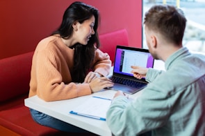 Two people are sitting at a table with a laptop, engaged in a discussion. The person on the right is pointing at the laptop screen, which displays some data or website interface. They are in a modern, well-lit environment with a red cushioned bench.