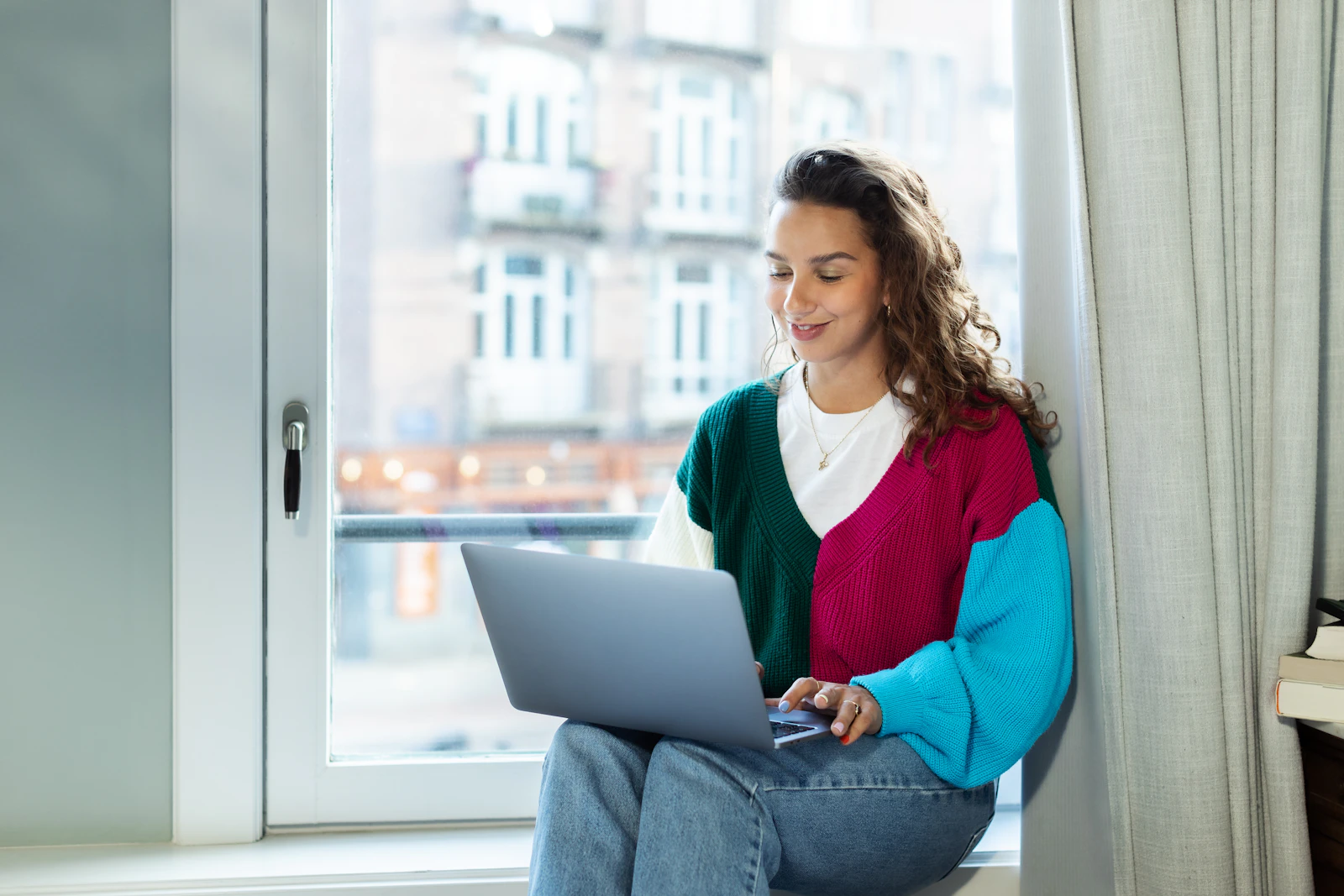 A solo freelancer sitting on a window sill working on a laptop in daylight