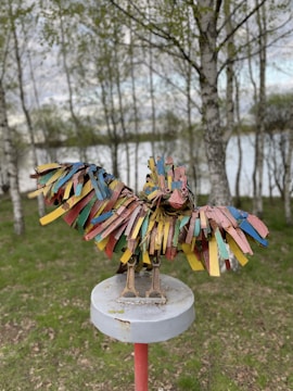 A colorful bird sculpture made from reclaimed metal and painted in vibrant hues of blue, red, yellow, and green. It stands on a round pedestal with a natural backdrop of trees and greenery.