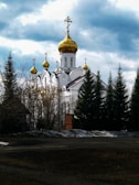 A picturesque scene of a white Orthodox church with distinctive gold domes surrounded by tall evergreen trees. The sky above is partly cloudy, adding depth to the landscape.