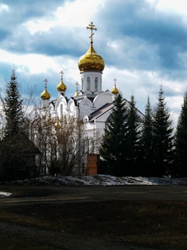 A picturesque scene of a white Orthodox church with distinctive gold domes surrounded by tall evergreen trees. The sky above is partly cloudy, adding depth to the landscape.