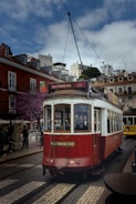 A classic red and white tram is moving down a cobblestone street lined with picturesque buildings. The tram is part of a city tour service, as indicated by the 'HILLS TRAMCAR TOUR' sign. The sky is partly cloudy, and there are a few people walking by on the street.