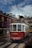 A classic red and white tram is moving down a cobblestone street lined with picturesque buildings. The tram is part of a city tour service, as indicated by the 'HILLS TRAMCAR TOUR' sign. The sky is partly cloudy, and there are a few people walking by on the street.