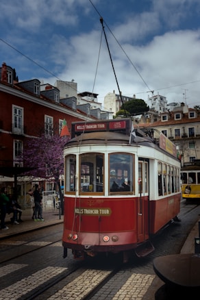 A classic red and white tram is moving down a cobblestone street lined with picturesque buildings. The tram is part of a city tour service, as indicated by the 'HILLS TRAMCAR TOUR' sign. The sky is partly cloudy, and there are a few people walking by on the street.