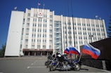 A large, multi-story government building with a beige facade, featuring flags and a government emblem at the top. Three motorcycles are parked in front, each adorned with the Russian flag. The sky is clear and blue, adding to the formal atmosphere.