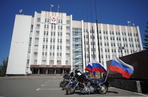 A large, multi-story government building with a beige facade, featuring flags and a government emblem at the top. Three motorcycles are parked in front, each adorned with the Russian flag. The sky is clear and blue, adding to the formal atmosphere.