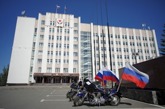 A large, multi-story government building with a beige facade, featuring flags and a government emblem at the top. Three motorcycles are parked in front, each adorned with the Russian flag. The sky is clear and blue, adding to the formal atmosphere.