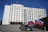 A large, multi-story government building with a beige facade, featuring flags and a government emblem at the top. Three motorcycles are parked in front, each adorned with the Russian flag. The sky is clear and blue, adding to the formal atmosphere.