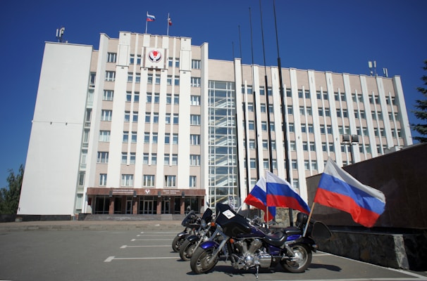 A large, multi-story government building with a beige facade, featuring flags and a government emblem at the top. Three motorcycles are parked in front, each adorned with the Russian flag. The sky is clear and blue, adding to the formal atmosphere.