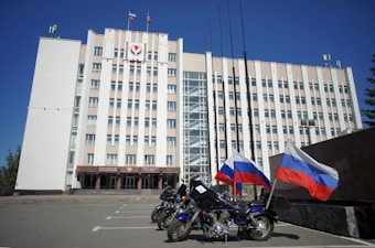 A large, multi-story government building with a beige facade, featuring flags and a government emblem at the top. Three motorcycles are parked in front, each adorned with the Russian flag. The sky is clear and blue, adding to the formal atmosphere.