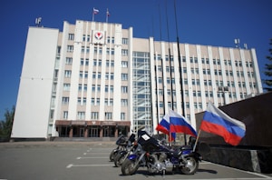 A large, multi-story government building with a beige facade, featuring flags and a government emblem at the top. Three motorcycles are parked in front, each adorned with the Russian flag. The sky is clear and blue, adding to the formal atmosphere.