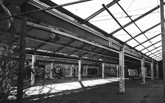 A large, abandoned industrial warehouse with a partially collapsed roof structure. Walls are covered with colorful graffiti, showcasing various artistic styles. The scene is devoid of people, and vegetation is encroaching through the open spaces.