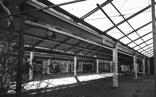 A large, abandoned industrial warehouse with a partially collapsed roof structure. Walls are covered with colorful graffiti, showcasing various artistic styles. The scene is devoid of people, and vegetation is encroaching through the open spaces.