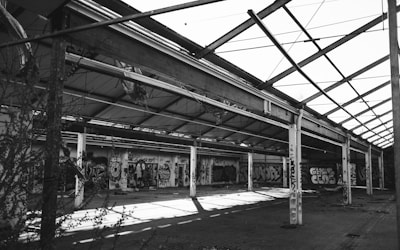 A large, abandoned industrial warehouse with a partially collapsed roof structure. Walls are covered with colorful graffiti, showcasing various artistic styles. The scene is devoid of people, and vegetation is encroaching through the open spaces.