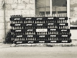 A stack of empty glass bottles organized neatly in black crates bearing the Pepsi logo and Arabic script. The crates are arranged in several rows and columns against a tiled building facade.